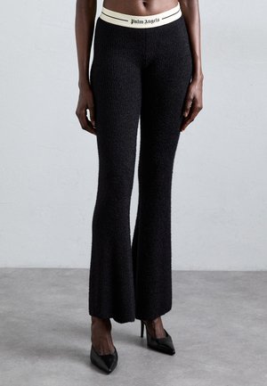 Mujer vistiendo pantalones acampanados negros texturizados, con una cinturilla blanca que lleva el logo de "Palm Angels", y tacones altos negros de punta, de pie sobre un suelo gris.