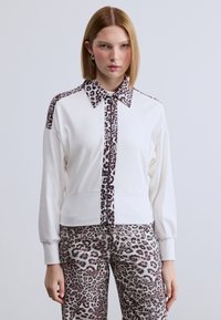 Λευκό μπουφάν με φερμουάρ και λεπτομέρειες λεοπάρ στο γιακά και τα μανίκια. Διαθέτει μακριά μανίκια και υφή στ ο ύφασμα. Συνδυάζεται με παντελόνια με το ίδιο μοτίβο λεοπάρ.