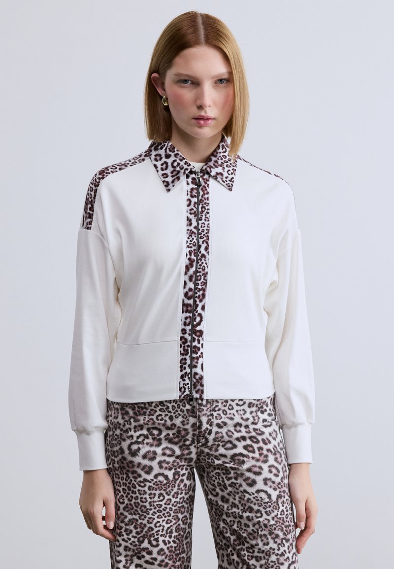 Λευκό μπουφάν με φερμουάρ και λεπτομέρειες λεοπάρ στο γιακά και τα μανίκια. Διαθέτει μακριά μανίκια και υφή στο ύφασμα. Συνδυάζεται με παντελόνια με το ίδιο μοτίβο λεοπάρ.