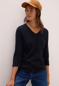 Marineblaues Langarmshirt mit V-Ausschnitt aus weichem Stoff, kombiniert mit hellblauen Jeans und einer sandfarbenen Wildledermütze, vor einem neutralen Hintergrund präsentiert.