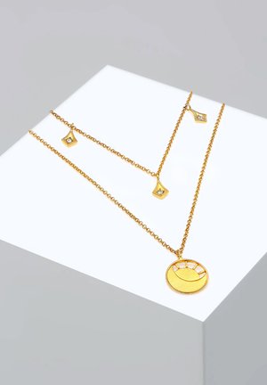 Un collier à plusieurs couches de couleur or présente un pendentif rond avec un motif de soleil et des accentuations géométriques, monté sur une fine chaîne en or ornée de petites embellissements.