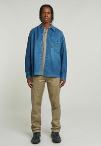 Niebieska koszula dżinsowa z kieszonką, szara koszulka polo pod spodem, beżowe spodnie i czarne trampki. Model stoi na neutralnym tle.