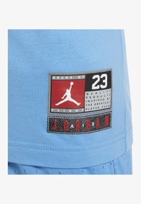 Světle modré bavlněné tričko s našitou nášivkou, která obsahuje černý, bílý a červený design, logo a textové detaily.