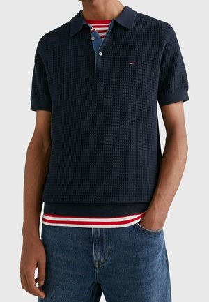 Marineblauwe gebreide polo met een gestructureerd patroon, korte mouwen en een kraag; gelaagd met een onderhemd met rode en witte strepen.