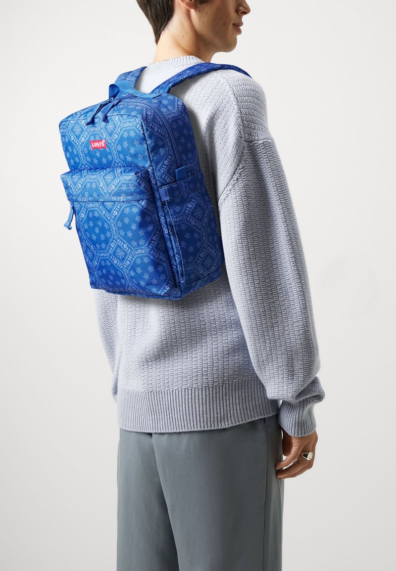 Personne portant un sac à dos Levi's à motifs bleus sur un pull en maille gris clair et un pantalon gris, se tenant devant un fond uni.