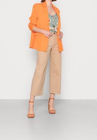 Blazer arancione, top a fantasia floreale, pantaloni beige a gamba larga e sandali con tacco arancioni. Tessuto liscio e design strutturato.