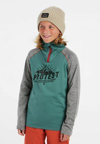 Pullover a maniche lunghe verde e grigio con colletto a zip, decorato con un logo di montagna, indossato con un berretto in maglia beige.