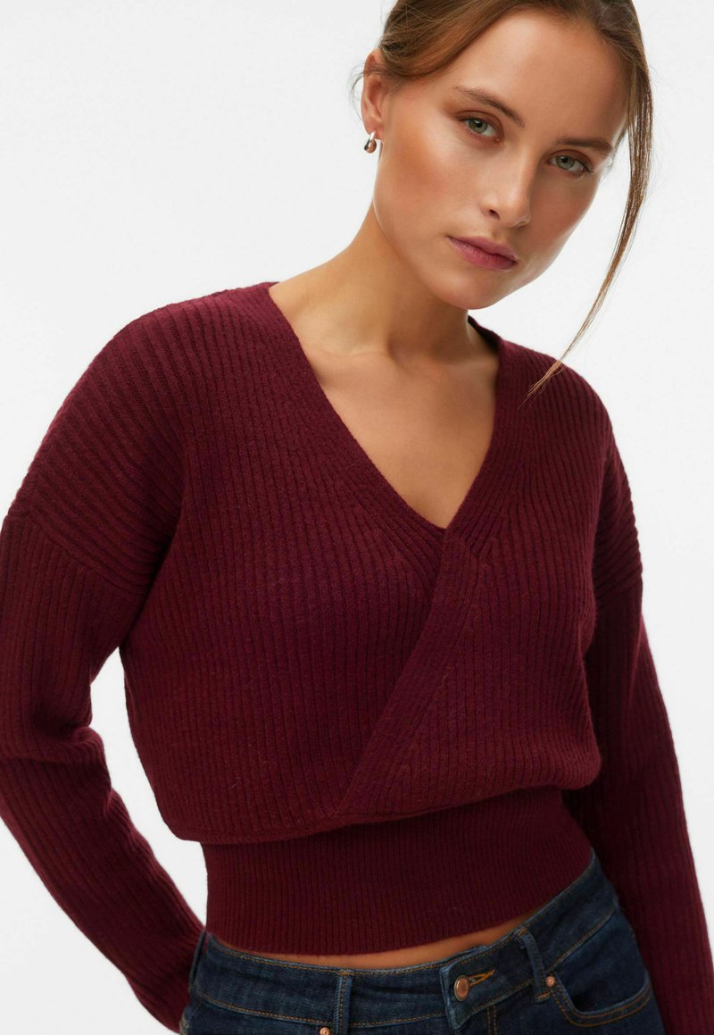 Femme portant un pull ajusté côtelé bordeaux à col en V avec manches longues et un jean en denim bleu foncé sur un fond blanc uni.