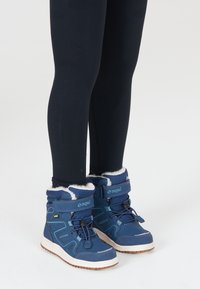 Blå vandtætte støvler med hvid fleece-foring, struktureret materiale, justerbare stropper og gummisål. Båret med sorte leggings.