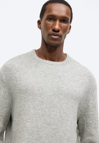 Ανοιχτό γκρι, πλεκτό πουλόβερ με στρογγυλή λαιμόκοψη και μακριά μανίκια, με απαλό υφή και μινιμαλιστικό σχεδιασμό. Χωρίς ορατά σχέδια ή διακοσμήσεις.