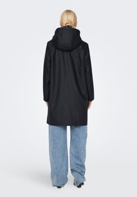 Manteau long noir imperméable avec capuche, présentant une coupe décontractée et une texture lisse. Associé à un jean large bleu clair.