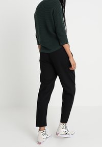 Pantalones negros ajustados combinados con zapatillas deportivas altas blancas. La parte superior es de un diseño suelto en verde oscuro, texturizado y con mangas de tres cuartos.