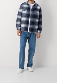 Giacca di flanella a quadri blu navy e bianchi con due tasche sul petto, chiusura a bottoni e colletto, abbinata a jeans in denim blu chiaro.
