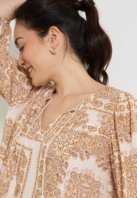 Blusa con estampado que presenta un fondo rosa con intrincados diseños de paisley y flores en dorado, un escote en V y una tela suave y ligera.