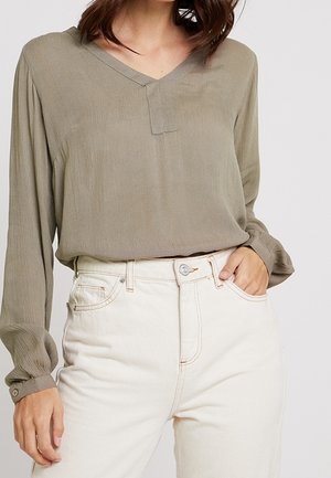 Frau trägt eine lockere, olivgrüne Langarmbluse, die in eine hoch taillierte, elfenbeinweiße Jeans gesteckt ist, mit einer Hand in der Tasche.