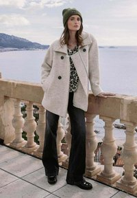 Abrigo de lana gris claro con botones negros, combinado con una blusa de estampado de leopardo, pantalones negros acampanados y un gorro de punto verde. Entorno al aire libre.