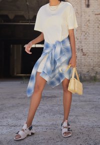 Vit kortärmad skjorta, ljusblå rutiga skjorta knuten runt midjan, beige sandaler, gul vadderad handväska, stående på en texturerad yta.