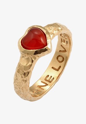 Auksinis žiedas su tekstūruotu paviršiumi, puikuojantis širdies formos raudonu akmeniu. Viduje išgraviruoti žodžiai "ŠVIESK MEILĘ."