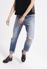 Vaaleansiniset revityt farkut, joissa on rento leikkaus, käännettävät lahkeet, valmistettu denimistä. Yhdistetty mustiin nauhoitettuihin kenkiin ja mustaan puseroon.