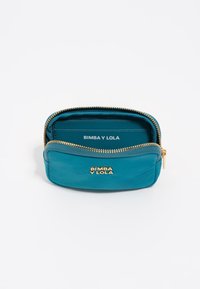 Bolso de cuero azul verdoso con cremallera y hardware dorado, que cuenta con un logo grabado y compartimentos para tarjetas en el interior. Textura suave y forma ovalada compacta.