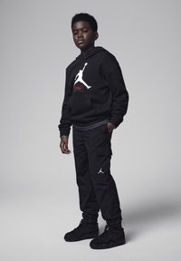 Haut à capuche noir avec logo Jumpman blanc, pantalon cargo noir avec poches, et chaussures noires avec accents de logo. Vue en pied sur fond gris.