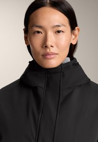 Veste noire avec un col montant, texture lisse et fermeture éclair. Elle présente un intérieur gris et des cordons réglables au niveau du col.