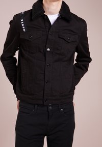 Giacca di denim nera con colletto nero testurizzato, chiusura frontale con bottoni e due tasche sul petto. Dettaglio logo bianco sulla spalla.