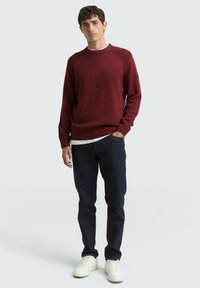 Granatroter Strickpullover mit gerippten Bündchen und Saum, kombiniert mit dunklen Jeans und weißen Turnschuhen. Einfaches Rundhalsdesign, glatte Textur.