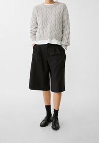 Helles graues, kabelgestricktes Sweatshirt mit gesenkten Schultern, über einem weißen Hemd getragen. Schwarze, maßgeschneiderte Shorts bis zur Mitte des Oberschenkels mit Falten und seitlichen Taschen.