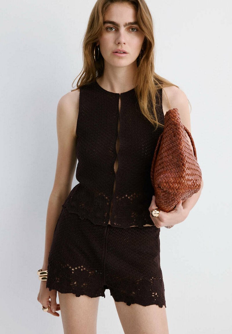 Femme portant un haut en maille noir sans manches et un short assorti, tenant une grande pochette tissée marron sur fond blanc.