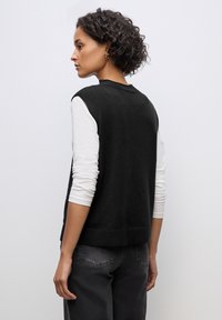 Maglione senza maniche nero in tessuto morbido, indossato sopra una camicia bianca a maniche lunghe. Il modello è in profilo, mostrando i capelli lunghi fino alle spalle e jeans scuri.