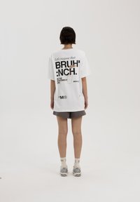 Witte oversized t-shirt met zwarte tekst en een afbeelding op de achterkant. Zachte katoenen stof, korte mouwen, casual ontwerp. Gepaarde met grijze shorts.