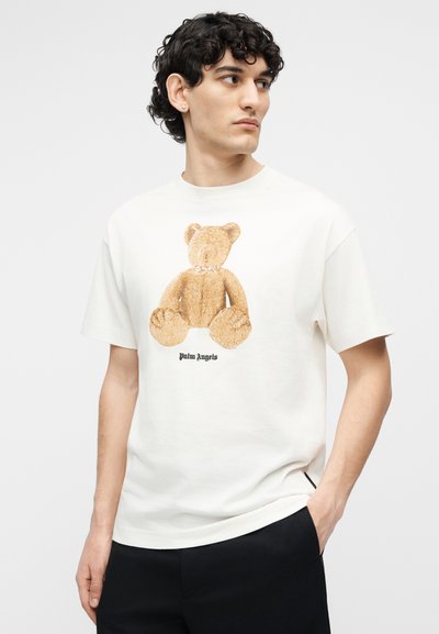 Jeune homme portant un t-shirt blanc avec un graphisme d'ours en peluche marron et le texte « Palm Angels », debout avec une main dans la poche.