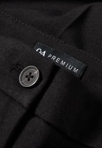 Svart tyg med en knapp och en synlig vittryckt "C&A PREMIUM" etikett sydd i en söm på plagget.