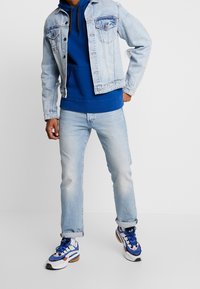 Light blue denim jacket with faded details, worn over a blue hoodie. Light blue jeans and white sneakers with blue and orange accents.