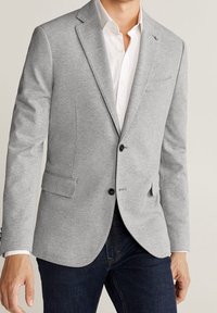 Grijze blazer van gestructureerde stof met een reverskraag, twee voorzakken en een enkele knoopsluiting, gedragen over een wit overhemd.