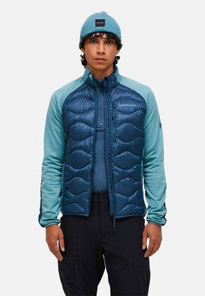 Blau gepolsterte Jacke mit türkisfarbenen Ärmeln, hohe Kragen, Reißverschluss und strukturierter Oberfläche. Getragen mit einer passenden Strickmütze.