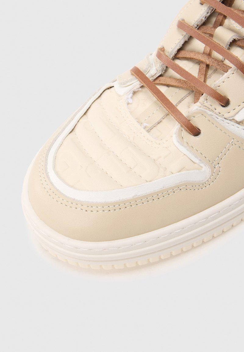 Beige sneaker featuring a textured leather upper, white stitching accents, and brown laces. Rounded toe and rubber sole for traction.