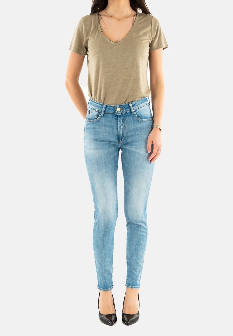 Comprador que lleva una camiseta de pico en verde oliva y jeans ajustados de color azul claro, combinados con zapatos de tacón de punta negra.