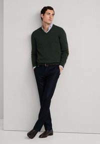 Tamnozeleni pulover s v-neck izrezom, ispod njega plava i bijela prugasta košulja, tamnoplave hlače, tamnosmeđe cipele, naslonjen na svjetlosivu zid.