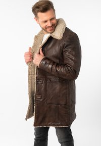 Abrigo de piel marrón con cuello de borrego, que presenta una superficie texturada, bolsillos frontales y cierres de botón, diseñado para proporcionar calidez y comodidad.