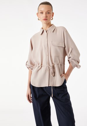 Woman wearing a beige button-up blouse with rolled sleeves and drawstring waist, paired with navy blue pinstripe pants.