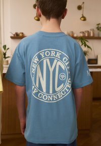 Lyseblå bomulds t-shirt med en cirkulær hvid grafik med teksten "NYC" og "Stay Connected" på bagsiden; korte ærmer, afslappet pasform.