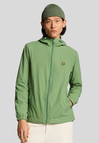 Chaqueta verde claro con cremallera y capucha, con puños elásticos, bolsillos laterales y un pequeño logo amarillo en el pecho. Textura lisa.