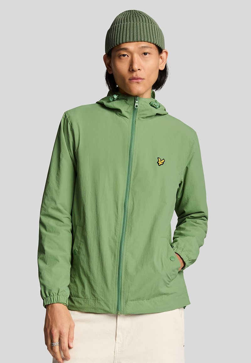 Chaqueta verde claro con cremallera y capucha, con puños elásticos, bolsillos laterales y un pequeño logo amarillo en el pecho. Textura lisa.