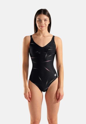 Giovane donna in piedi che indossa un costume intero nero con sottili motivi a strisce riflettenti e un piccolo logo bianco sull'anca.