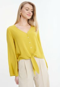 Blusa gialla a maniche lunghe con scollatura a V e dettaglio annodato sul davanti. Presenta piccoli bottoni e un tessuto morbido e testurizzato. Indossata con pantaloni beige.