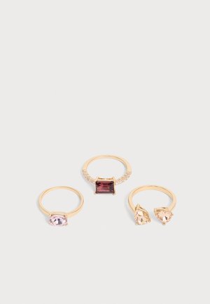 Trois bagues en or avec différentes pierres précieuses : une avec une gemme rectangulaire sombre, une avec une gemme ovale rose clair, et une avec deux gemmes en forme de goutte couleur pêche.