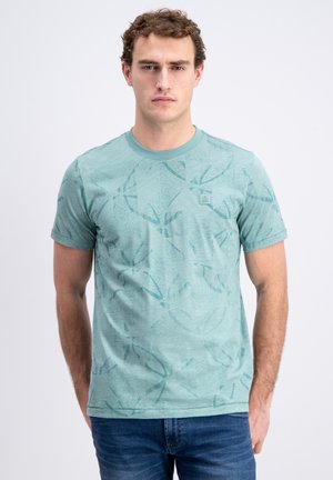Man draagt een licht turquoise T-shirt met patroon en korte mouwen en een blauwe spijkerbroek, staand tegen een effen witte achtergrond.