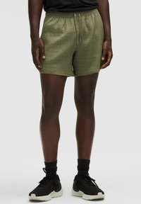 Olīvzaļi sporta šorti ar teksturētu virsmu, elastīgu jostu un auklu. Apvienoti ar melniem sporta apaviem un zeķēm.